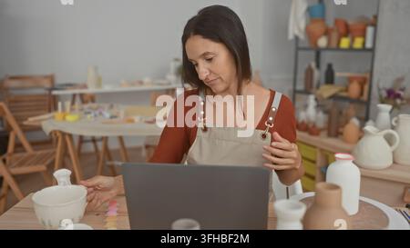 Woman potter holds a ceramic mug with her hand at a studio table beside a laptop, inspecting glaze and rotating the piece while seated; quiet concentr Stock Photo