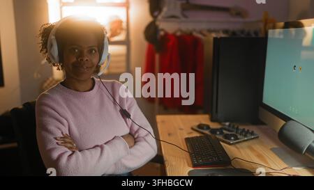 Young african american woman wearing white headphones typing on keyboard at wooden desk with monitor and audio mixer in studio; focus remote work. Stock Photo