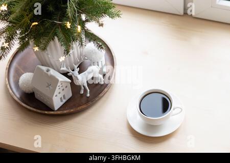 Black coffee in a white cup beside evergreen branches, ceramic house and reindeer on a wooden windowsill, with large copy space for Christmas design. Stock Photo