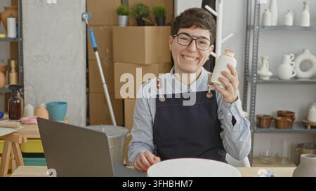 Woman holding ceramic vase on a turntable in a studio while wearing an apron and working on a laptop; craft business creativity. Stock Photo