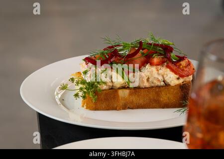 Smoked fish salad on toasted bread, generously topped with fresh tomato, red onion, spring onion, dill and parsley, served on a white plate. Stock Photo