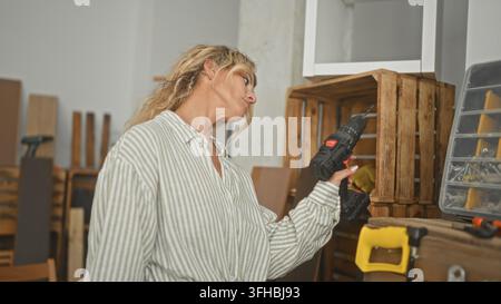 Woman holding drill inspects drill bit while standing by wooden crate in studio; concentration craft skill. Stock Photo
