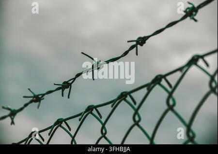 Barbed wire atop chain-link fence forms a stark boundary against an overcast sky, evoking security, restriction, confinement and a tense sense of barr Stock Photo