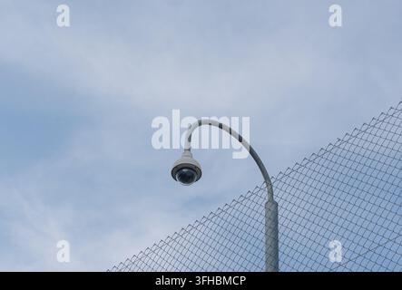 Low-angle shot of a surveillance security camera mounted on a curved pole with a diagonal wire mesh fence and a soft daylight sky background Stock Photo