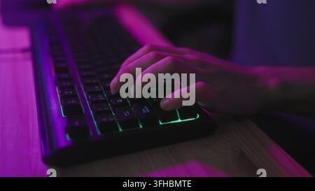 Young hispanic man typing with hands on rgb backlit gaming keyboard in studio at wooden desk under purple neon lighting; focused immersion. Stock Photo
