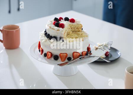 Two-tier white-frosted cake is sitting on pedestal stand on glossy counter, showing berry topping Stock Photo