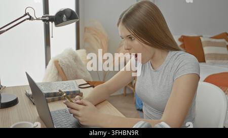 Woman holds smartphone and laptop while smiling and fist pump at desk in bedroom; study success joy. Stock Photo