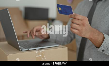 Young indian man holds creditcard in one hand and types on laptop with the other amid moving boxes in a building; convenience. Stock Photo