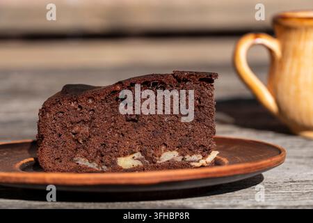 Chocolate cake slice on plate, close up. Slice of homemade chocolate brownie cake with apples on clay plate, with ceramic cup blurred in the backgroun Stock Photo