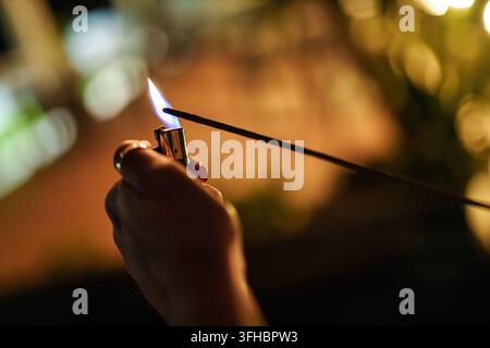 Woman's hand holding a lighter with a bright flame, igniting a long, thin sparkler stick, creating a warm glow in a dark outdoor setting with bokeh li Stock Photo