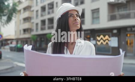 Woman architect in hardhat holds blueprints and looks up on a city street in front of a building; determination planning focus. Stock Photo