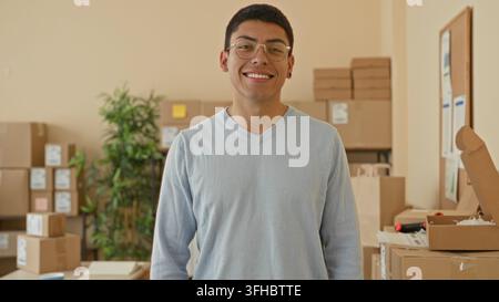 Man smiles beside stacked cardboard boxes and an open parcel on a table in a building packing room; cheerful entrepreneurship. Stock Photo