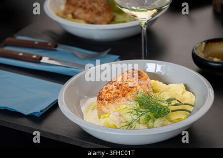 Golden-brown fish patty served with a fresh green salad and aromatic dill, presented in a white bowl on an elegant dark dining table. Stock Photo