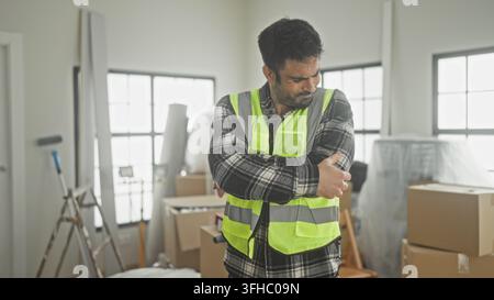 Man clutching his left arm and wincing next to packed boxes, ladder and paint roller during interior renovation in a building; safety concern. Stock Photo