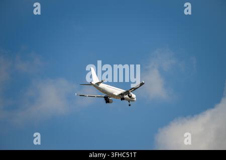A white passenger airplane descends through a blue sky with soft white clouds, preparing for landing with its gear extended. Stock Photo