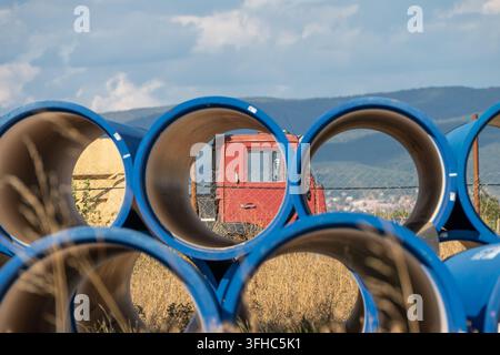 A stack of large blue industrial water pipes frames a red vintage work truck parked in a dry rural field against a backdrop of distant hills and cloud Stock Photo