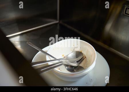 Dirty bowl, plate and cutlery with food residue sit inside stainless steel kitchen sink. Concept of dishwashing and kitchen cleanup Stock Photo