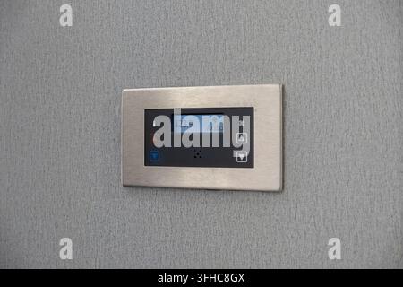 The brushed metal electronic thermostat unit sits on a textured grey wall, displaying current heating and cooling settings to manage comfort levels in Stock Photo