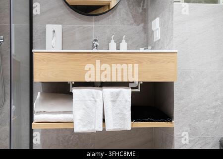 Minimalist bathroom sink unit featuring natural wood veneer shelving with neatly folded white towels and soap containers, providing a functional desig Stock Photo