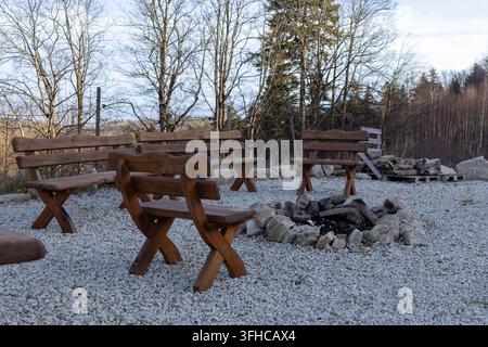 Empty rustic wooden benches are arranged around a stone fire pit on a gravel ground, promoting outdoor social gatherings, tranquil camping, and peacef Stock Photo