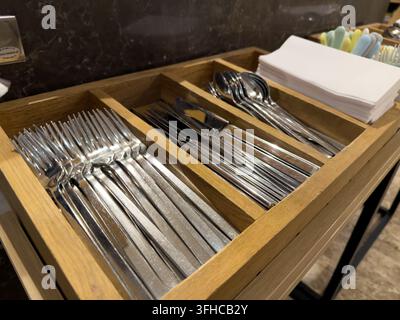 Detailed shot of sparkling clean stainless steel forks, knives, and spoons neatly arranged in a wooden holder, alongside crisp white napkins, for hygi Stock Photo