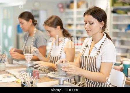 During working day, three employees of pottery workshop molding from clay, paint finished product. They apply pattern to surface of plate with brush, Stock Photo