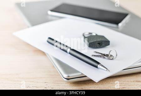 A closed padlock, two keys and a black pen lie on white paper on a closed silver laptop, with a dark phone behind. Physical lock as a metaphor for sec Stock Photo