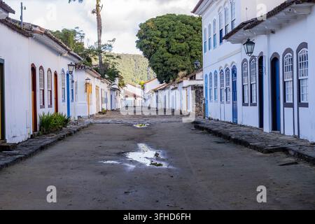 View of a quiet colonial street in Paraty, Brazil. White houses with colorful trims line the cobblestone path. Puddles reflect the sky on the ground. Stock Photo