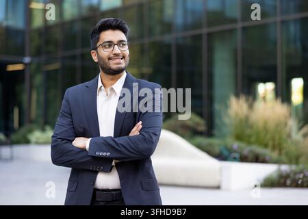 Confident young Indian businessman standing with arms crossed, professional corporate portrait of a successful entrepreneur in a modern office park. Successful career and leadership. Stock Photo