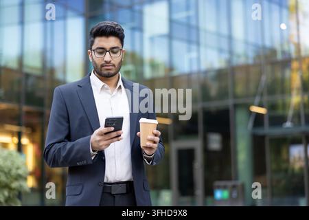 Professional businessman using smartphone and drinking coffee, remote work and digital nomad lifestyle in modern city office district. Corporate employee checking mobile device while commuting. Stock Photo
