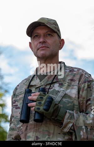 A man in camouflage uniform holds binoculars while looking upward outdoors. Stock Photo