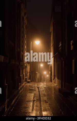 Atmospheric narrow alley Stradela Vantului with warm streetlights glowing in dense fog at night in Constanta old town, Romania⁠ Stock Photo