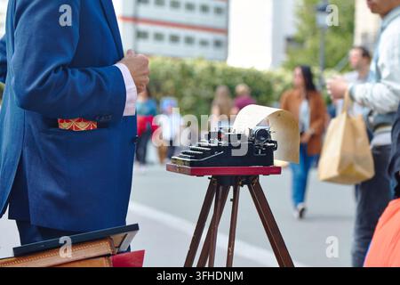 A close-up shot of an old mechanical typewriter with paper on a tripod, with a man in a blue suit standing nearby on a blurry city street sidewalk wit Stock Photo