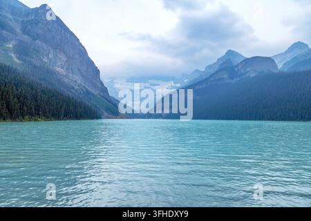A serene view of a vast, clear turquoise lake surrounded by towering, rugged mountains and lush green pine forests on a hazy day, 111 Lake Louise, AB Stock Photo
