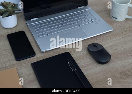 Top angle view of a clean, modern workspace featuring an open silver laptop, a black smartphone, a wireless mouse, a black notebook, and a pen on a li Stock Photo