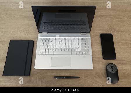 Overhead flat lay composition of a professional remote work desk featuring an open silver laptop, black notebook, pen, wireless mouse, and smartphone Stock Photo