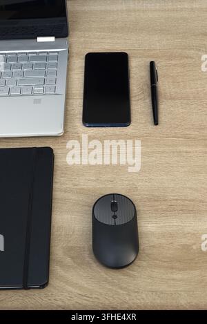 Top view geometric composition of office tools including a smartphone, pen, notebook corner, and wireless mouse arranged next to a laptop on a wooden Stock Photo
