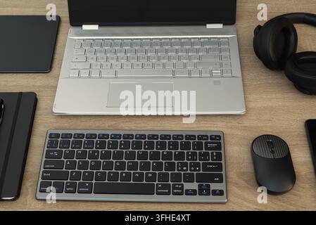 Overhead flat lay view of a fully equipped modern office desk featuring an open silver laptop, external compact keyboard, wireless headphones, tablet, Stock Photo
