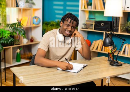 African American young man on home table writes notes in notebook with pen for study planning at home. Black guy on home table reviews written task list with happy focused mood in living room interior Stock Photo