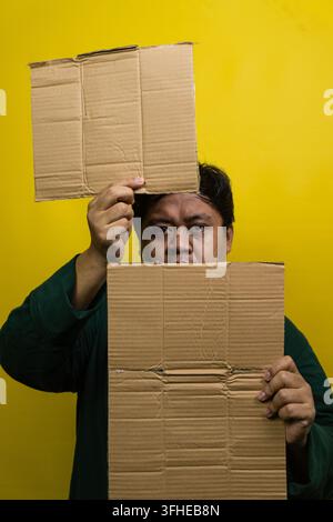 An Asian man holding two blank pieces of brown corrugated cardboard against a solid yellow background. One sign is held above the head, while the othe Stock Photo