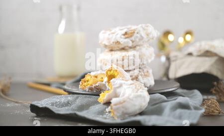 A stack of sweet glazed donuts sits on a dark plate, featuring a broken pastry piece showing its fluffy crumb texture in soft natural light. Stock Photo