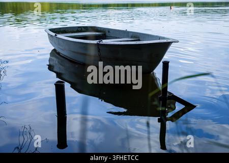 An empty metal rowboat moored on a calm forest lake, with reflections on the water and a distant swimmer blurred in the background. Peaceful summer sc Stock Photo