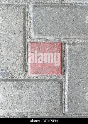 Detailed top-down view of a worn red paving block embedded among grey concrete pavers. The contrasting colour, rough surface and geometric arrangement Stock Photo