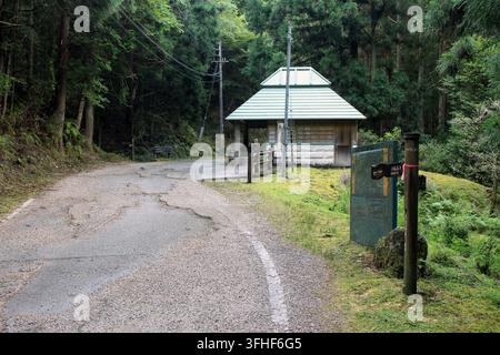 A small mountain shelter and rest stop with a tiered roof stands beside a cracked asphalt road within a dense cedar forest. Stock Photo