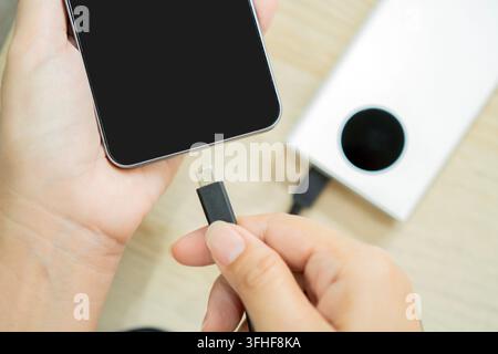 High angle view of a person's hands inserting a USB charging cable into a smartphone next to a portable power bank. Stock Photo