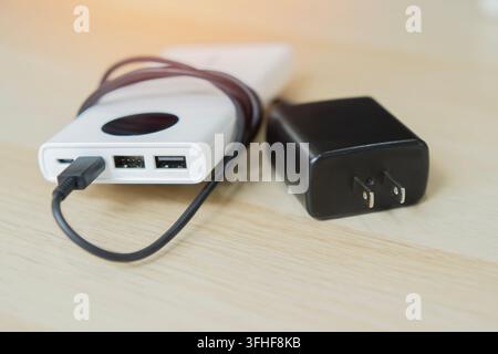 A white portable power bank connected with a USB charging cable resting next to a black wall adapter plug on wooden desk. Stock Photo