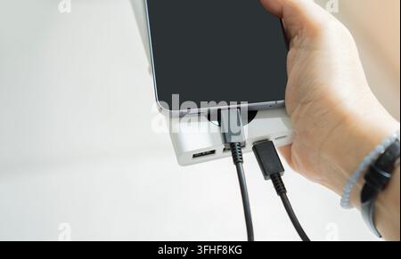 A person's hand holding a black smartphone connected to a white power bank via USB cable on a tabletop setup. Copy space. Stock Photo