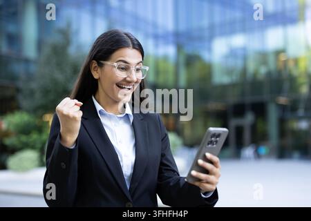 Career success and professional achievement, happy businesswoman celebrating good news on smartphone in modern office district. Excited female professional reacting to positive notification on mobile. Stock Photo