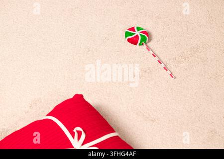 A decorative holiday candy swirl on a stick placed near a vibrant red present with a white ribbon on a cozy beige surface with ample copy space Stock Photo
