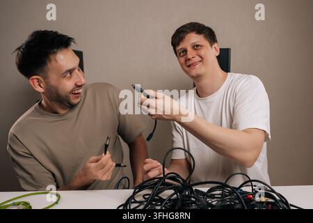 Two smiling men engaging with challenging mess of disorganized cables and cords, finding humor in the task of identifying different connectors and Stock Photo